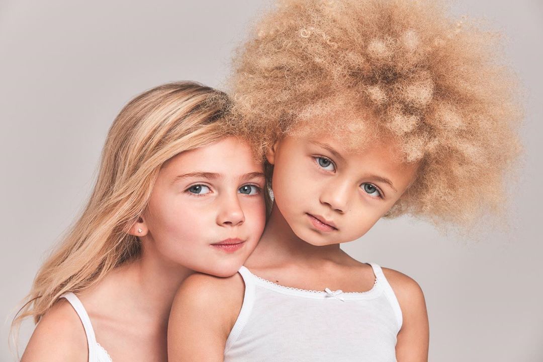 Two young girls, one with blonde hair and the other with curly afro-textured hair, pose closely together in a studio portrait.