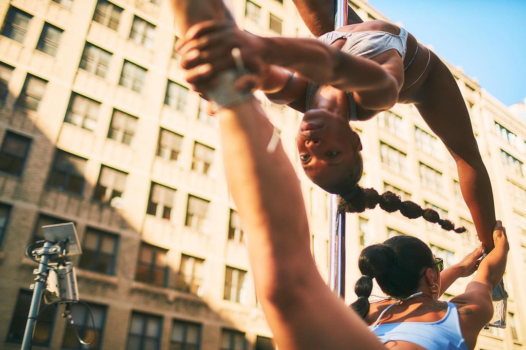 Two women perform pole dancing in an urban setting in daytime. Buildings are visible in the background. The women are wearing athletic apparel.