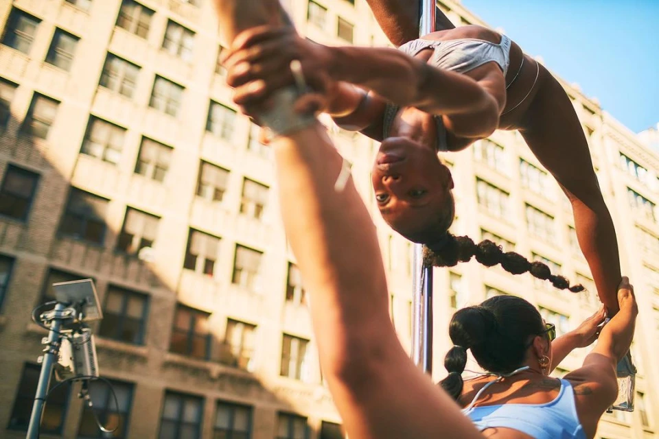 Two women perform pole dancing in an urban setting in daytime. Buildings are visible in the background. The women are wearing athletic apparel.
