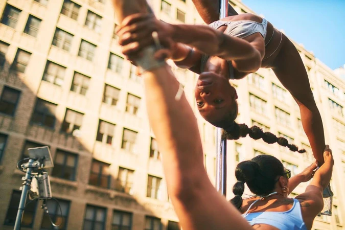 Two women perform pole dancing in an urban setting in daytime. Buildings are visible in the background. The women are wearing athletic apparel.