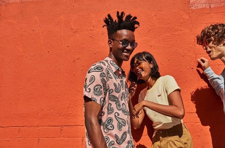 Three friends smile and laugh in front of a solid orange background, dressed in fashionable patterned shirts and trendy sunglasses.