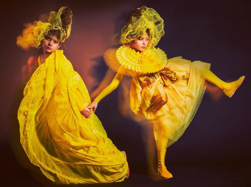 Two children holding hands in yellow haute couture dresses and headwear, captured with studio lighting against a muted background.