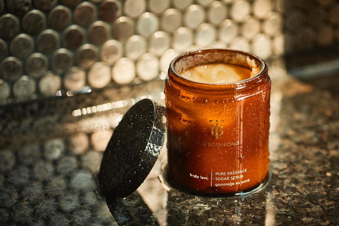 Amber glass jar of sugar scrub with lid on textured counter, soft lighting, and a mosaic tile wall. Focus on skincare and body care.