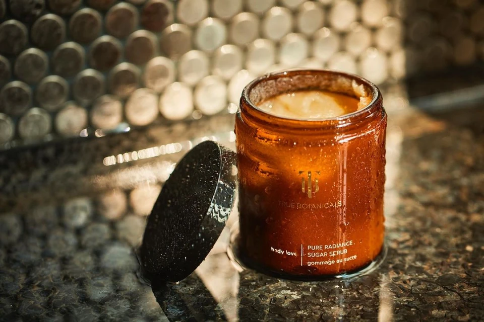 Amber glass jar of sugar scrub with lid on textured counter, soft lighting, and a mosaic tile wall. Focus on skincare and body care.