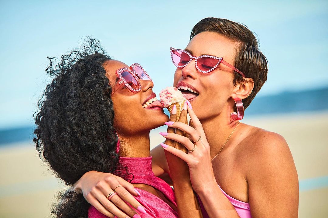 Two women share an ice cream cone on a sunny day, wearing pink clothing and sunglasses against a bright blue sky and neutral background.