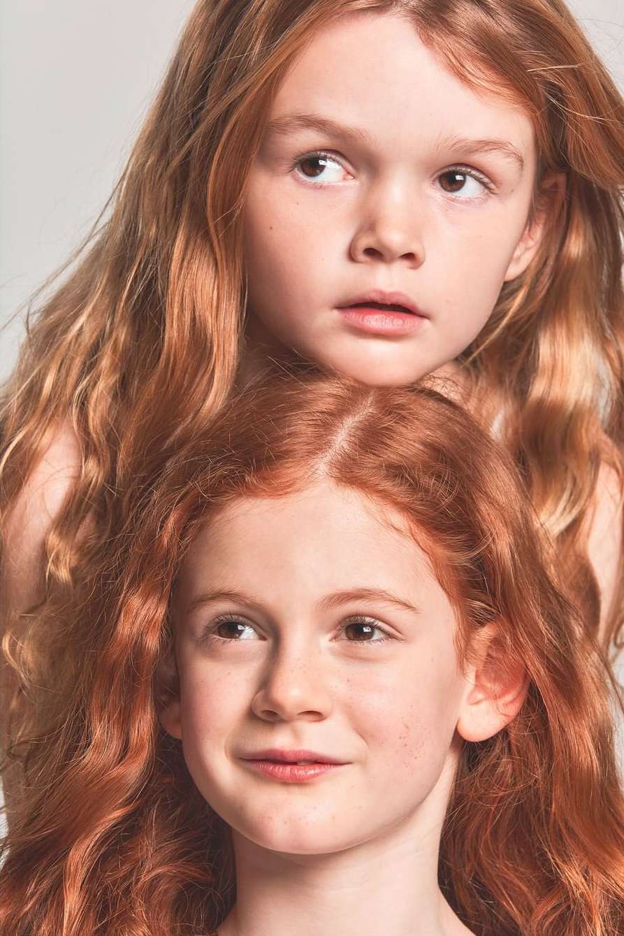 Portrait of two red-haired young girls, one slightly behind the other, posing in a studio environment with a neutral backdrop.