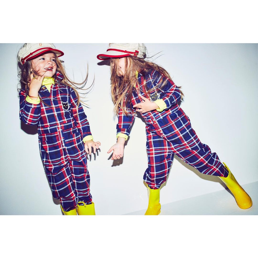 Two kids in plaid jumpsuits and matching hats pose playfully in a studio. The children wear bright yellow boots and light yellow turtlenecks.
