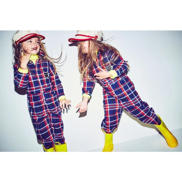 Two kids in plaid jumpsuits and matching hats pose playfully in a studio. The children wear bright yellow boots and light yellow turtlenecks.