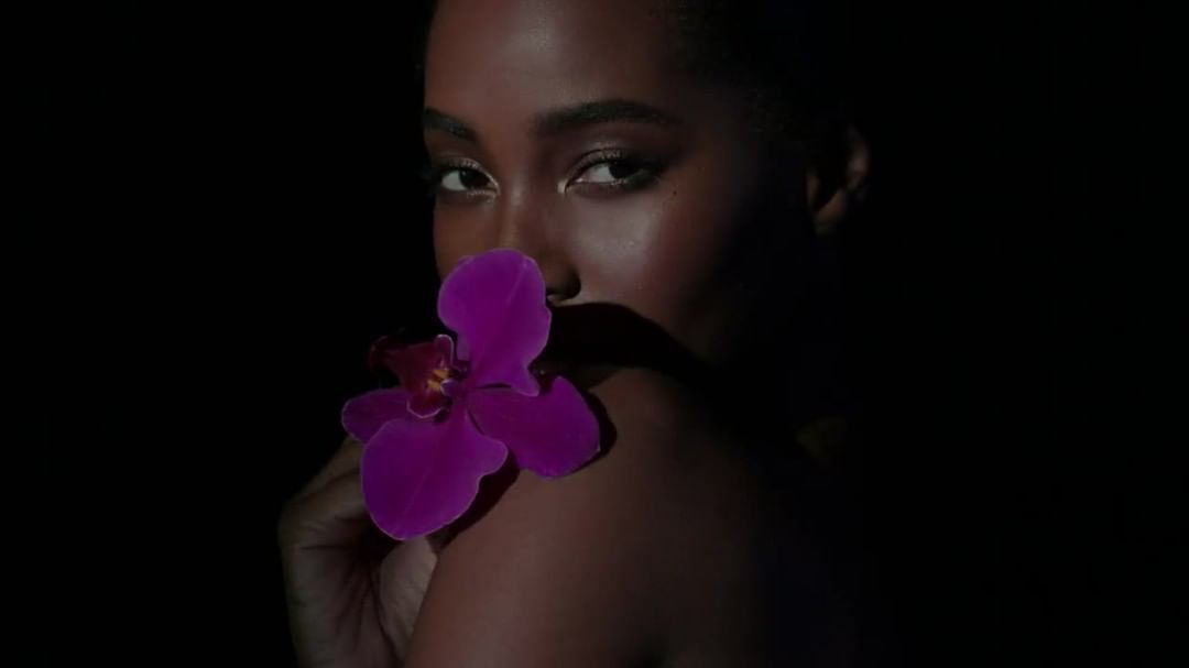 Close-up portrait of a woman with dark skin, dark eyes, and dark hair, holding a bright purple orchid flower against a dark background.
