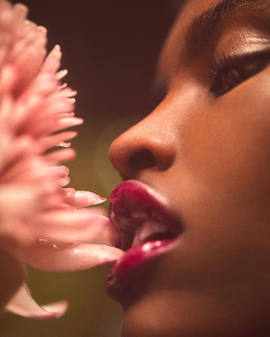 Close-up profile of a woman with dark skin and red lipstick, posing with a pink flower in a soft, filtered light setting.