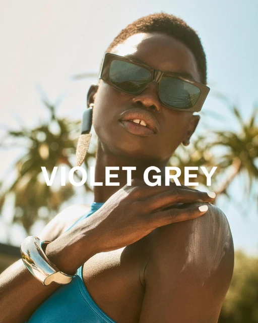 A dark-skinned woman models sunglasses, wearing a blue swimsuit and jewelry in an outdoor setting with palm trees and bright sunlight.