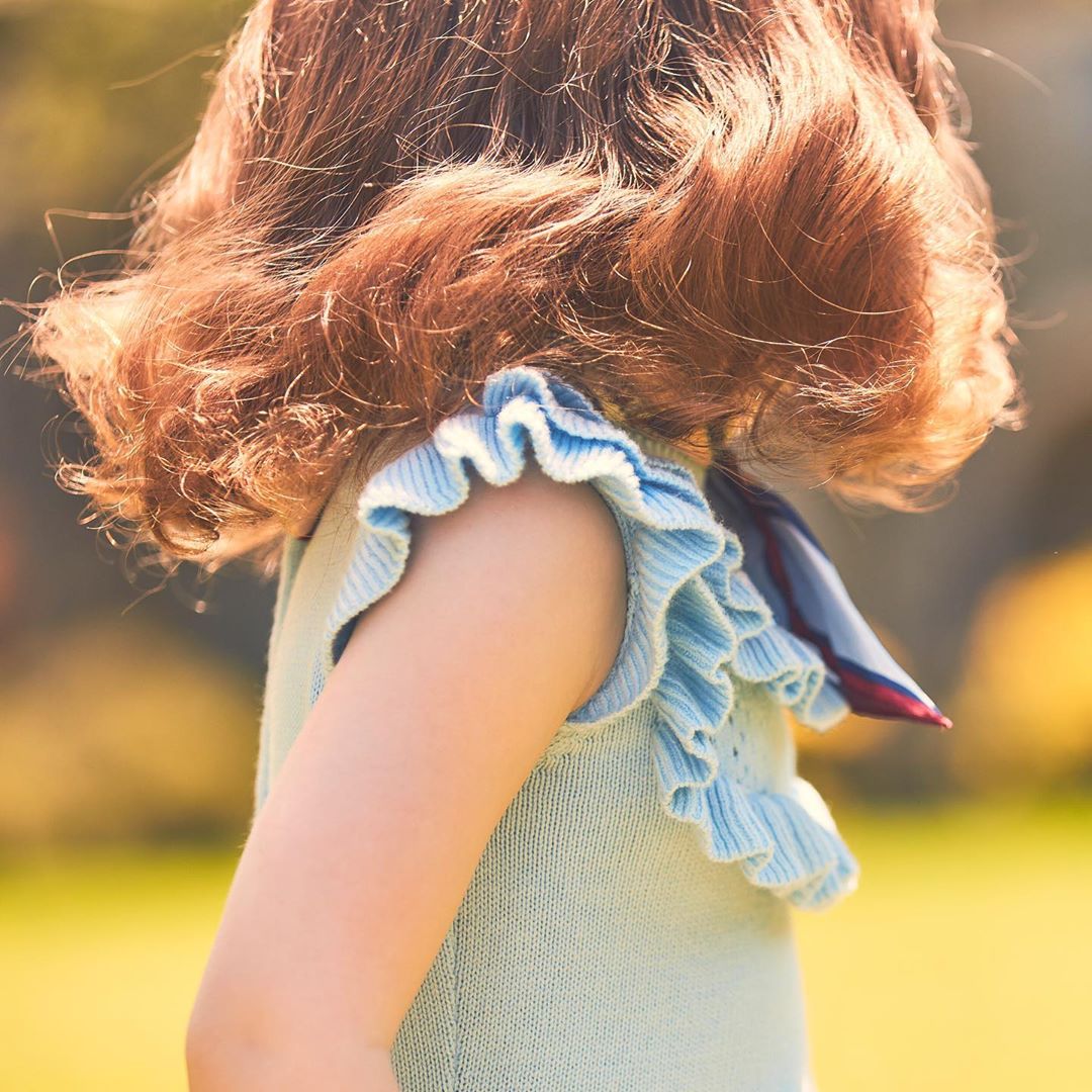 Child with auburn curly hair wearing a light blue dress with ruffles standing outside in the sun. Soft focus and shallow depth of field.