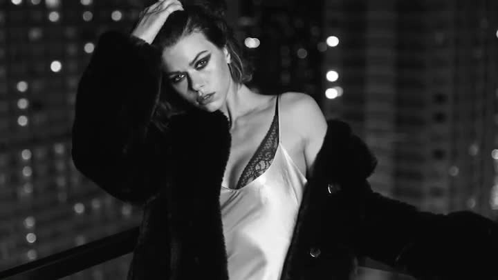 Black and white portrait of an adult female in a fur coat and lingerie top, standing indoors with a blurred city lights background.