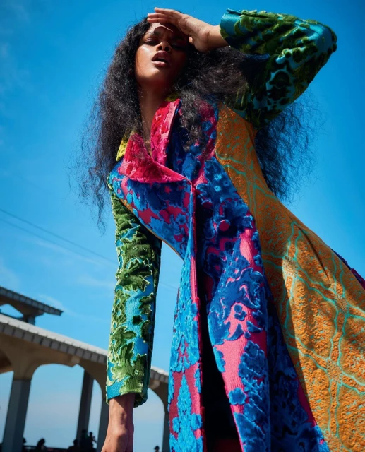 Stylish woman in a patterned coat poses under a blue sky with architectural elements in the background, creating a vibrant fashion image.