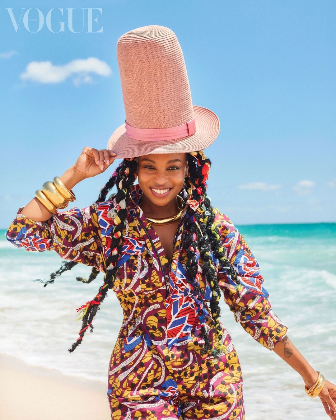 Woman on beach wearing tall straw hat and patterned jumpsuit, smiling brightly with ocean and blue sky background, jewelry accents.