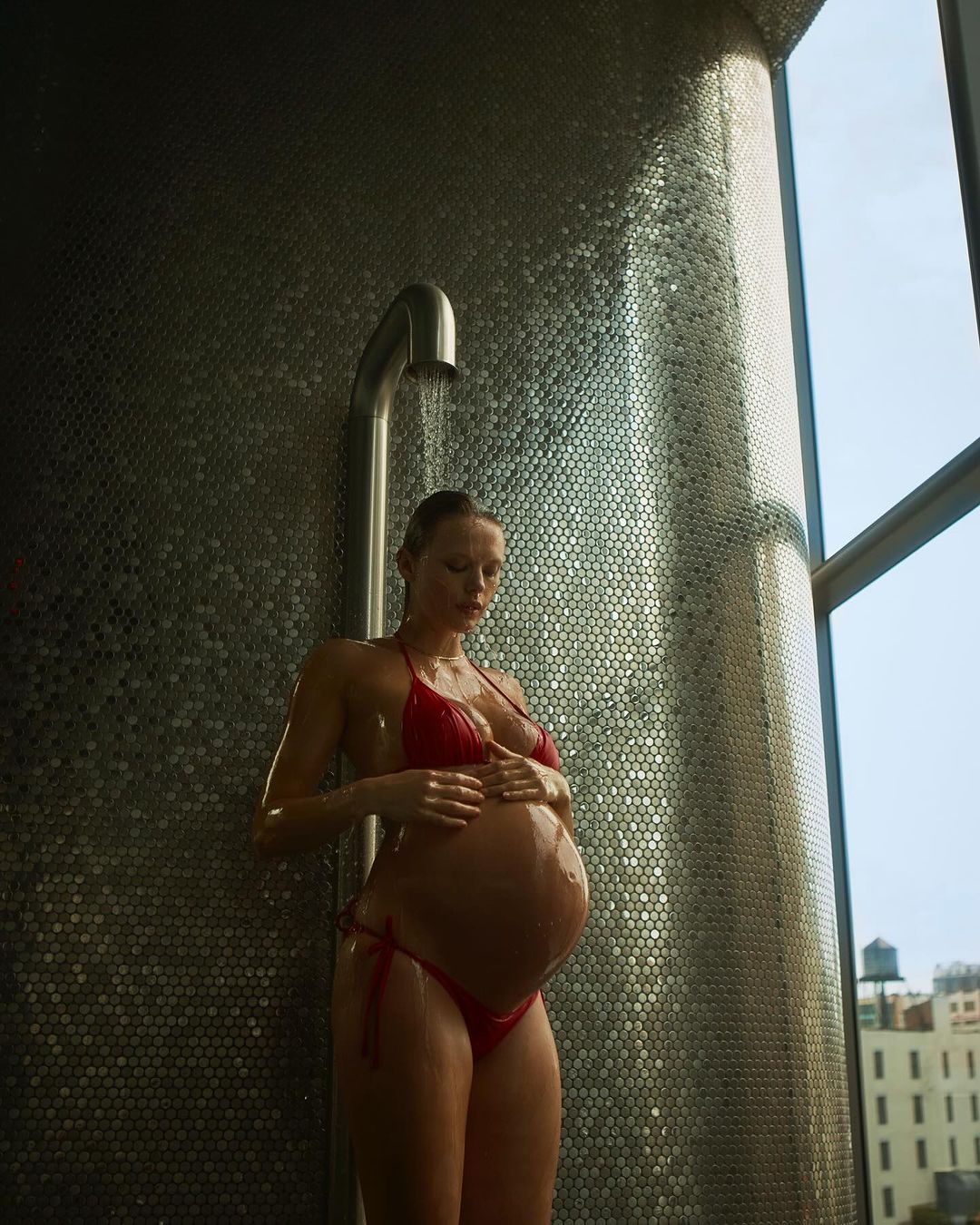 Pregnant woman in red bikini standing under shower. Silver tiled wall and bright window, natural light, clear water, wellness concept.