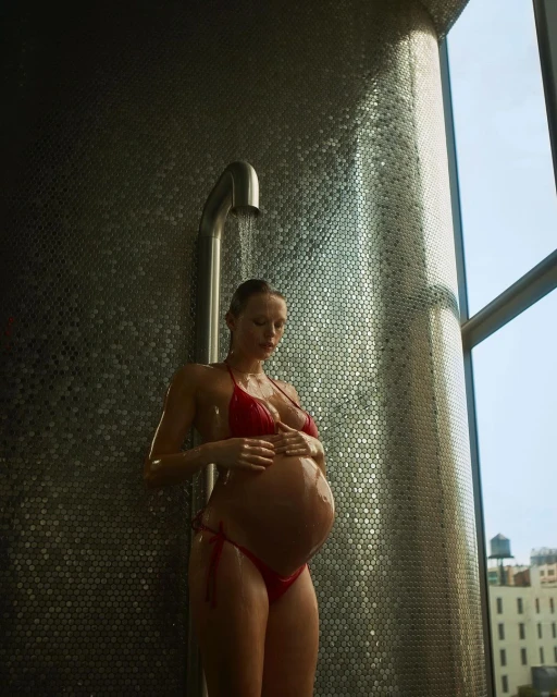 Pregnant woman in red bikini standing under shower. Silver tiled wall and bright window, natural light, clear water, wellness concept.