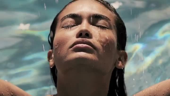 Close-up of an adult woman with her face turned up to catch the water droplets in an outdoor setting with a blue-toned background.