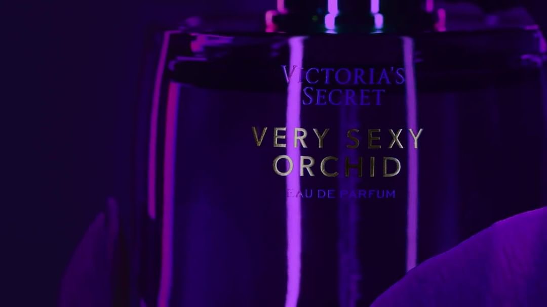 Close-up studio shot of a 'Victoria's Secret Very Sexy Orchid' perfume bottle against a vivid purple backdrop with vibrant lighting.