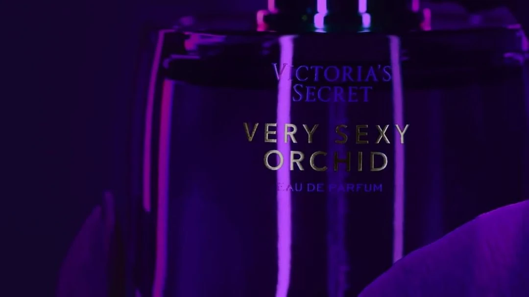 Close-up studio shot of a 'Victoria's Secret Very Sexy Orchid' perfume bottle against a vivid purple backdrop with vibrant lighting.