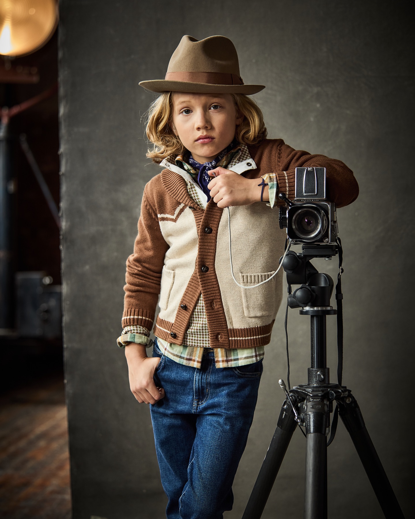 Boy with blond hair, wearing a hat and cardigan, poses next to a vintage camera on a tripod in a studio setting.