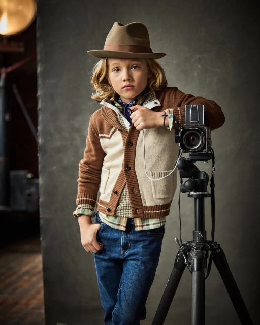 Boy with blond hair, wearing a hat and cardigan, poses next to a vintage camera on a tripod in a studio setting.