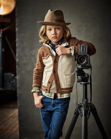 Boy with blond hair, wearing a hat and cardigan, poses next to a vintage camera on a tripod in a studio setting.