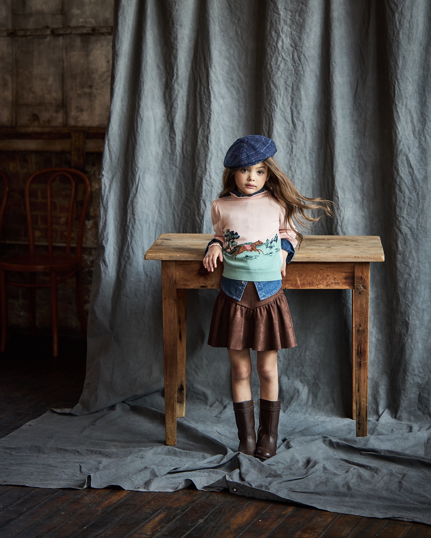 Young girl in stylish outfit with beret, sweater, skirt, and boots posing in front of a wooden table and grey backdrop.