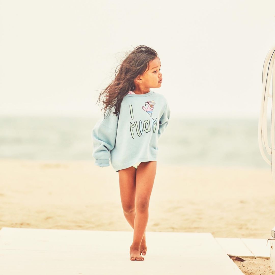 Young girl in a blue sweatshirt with text walks barefoot on a beach boardwalk, ocean and sky in background, medium shot, bright light.