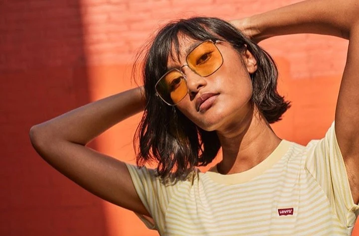 Portrait of a woman with orange sunglasses and a striped yellow top, posing in front of an orange wall with sunlight casting shadows.