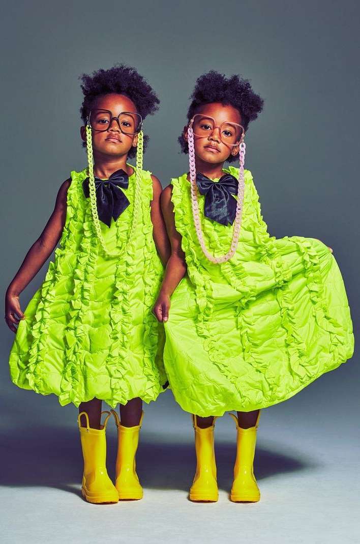 Two children stand side-by-side, dressed in matching bright green dresses, black bowties, and yellow rain boots against a grey background.