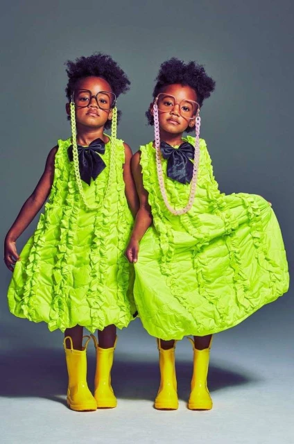 Two children stand side-by-side, dressed in matching bright green dresses, black bowties, and yellow rain boots against a grey background.
