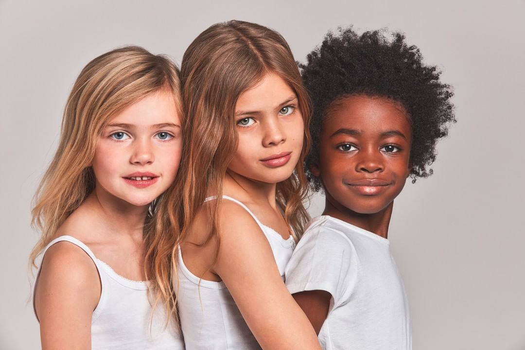 Close-up of three young girls of mixed ethnicities with blonde and brown hair, smiling in white tank tops against a grey background.
