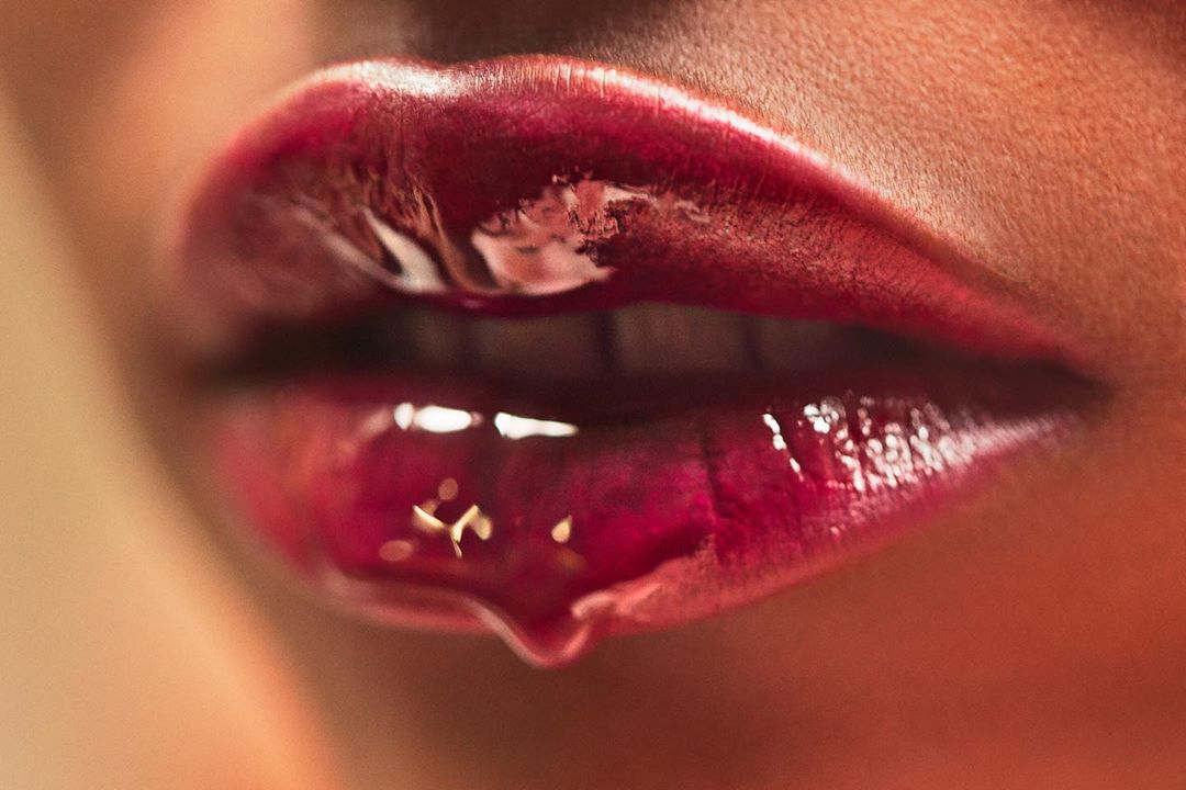 Extreme close-up of a woman's mouth with glossy burgundy lipstick, highlighted by a bright, soft light. A drip of gloss hangs below the lip.