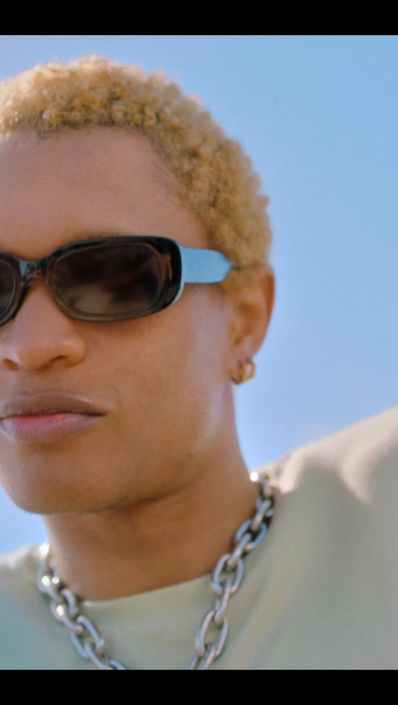 Close-up portrait of a person with blonde short hair wearing dark sunglasses and a silver chain necklace against a clear blue sky background.