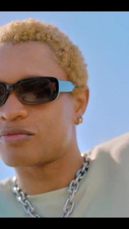 Close-up portrait of a person with blonde short hair wearing dark sunglasses and a silver chain necklace against a clear blue sky background.