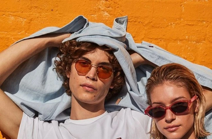 Two young adults wearing colored sunglasses pose against an orange brick wall, showcasing casual fashion and youthful style.