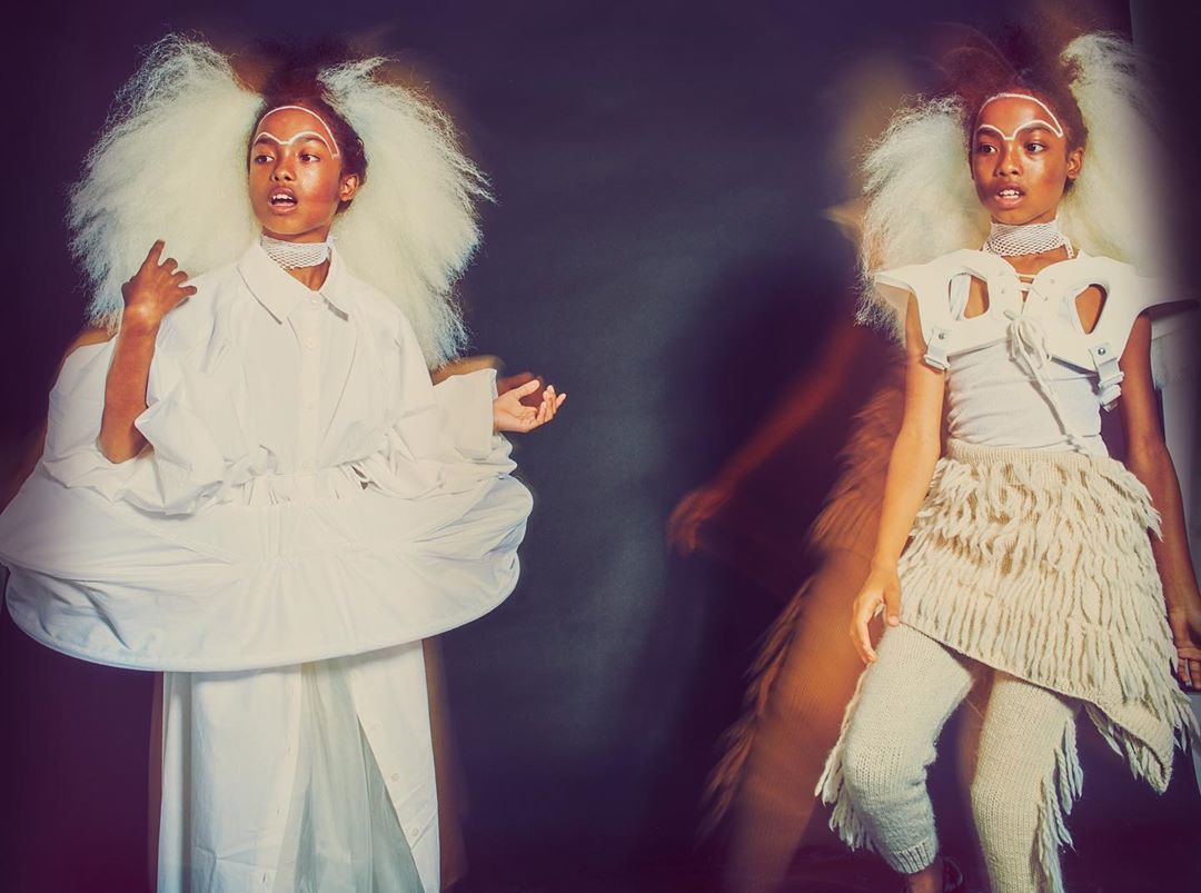 Two women pose in avant-garde fashion, one in a voluminous white dress, the other in knitted textures against a dark, moody backdrop.