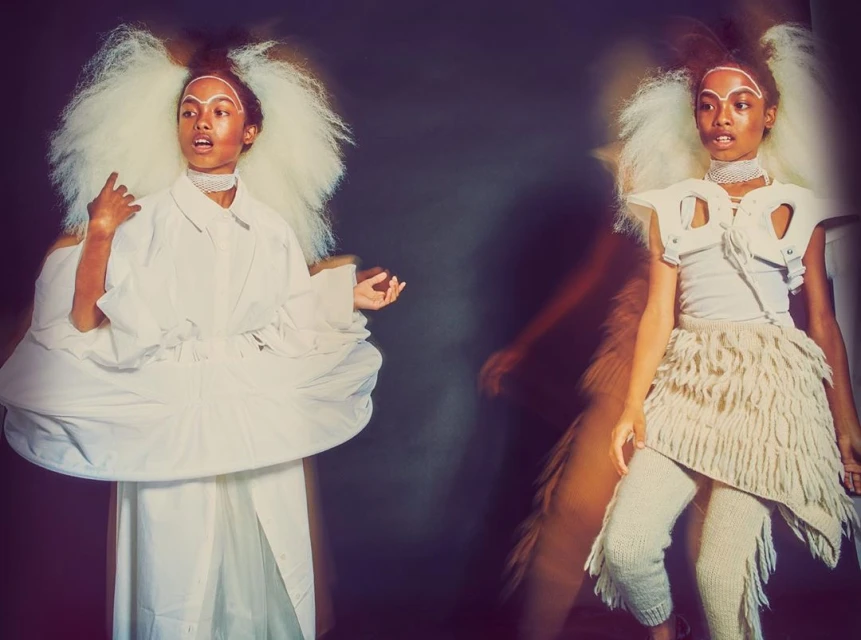 Two women pose in avant-garde fashion, one in a voluminous white dress, the other in knitted textures against a dark, moody backdrop.