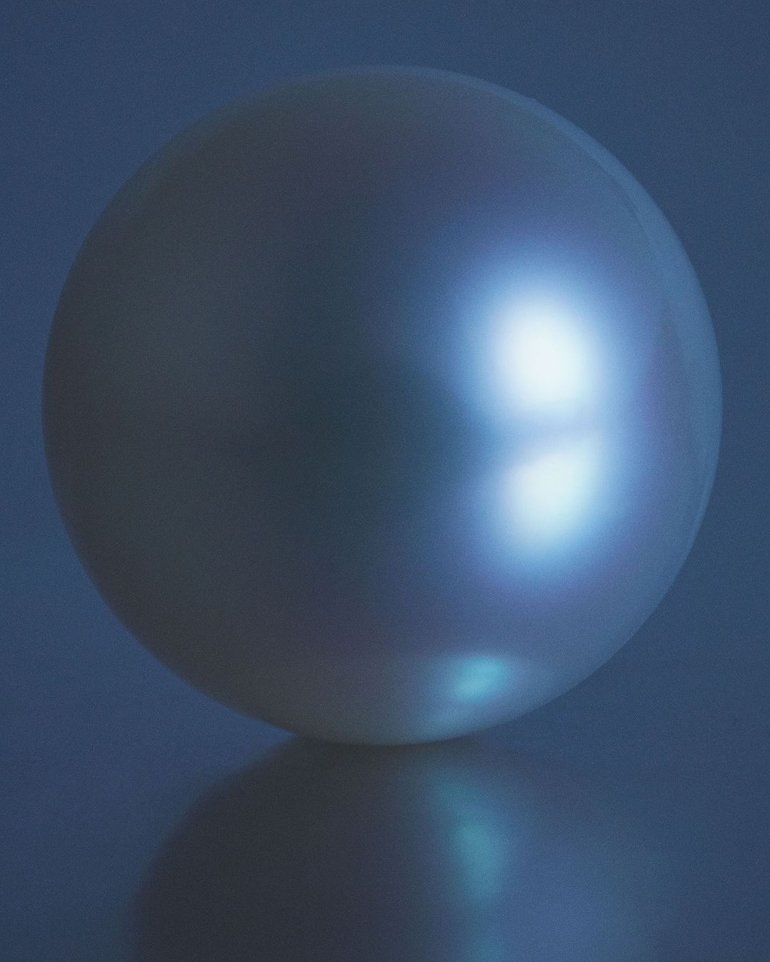 Close-up studio shot of a round, iridescent pearl against a dark blue background with a visible reflection.