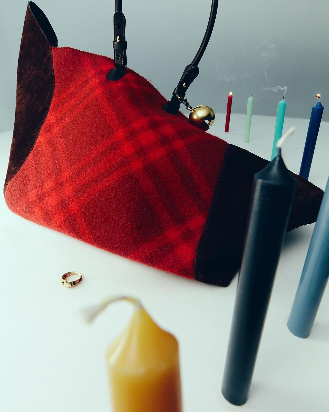 Still life studio shot of a plaid bag with a gold bell, a gold ring, and lit candles of different colors on a white surface.