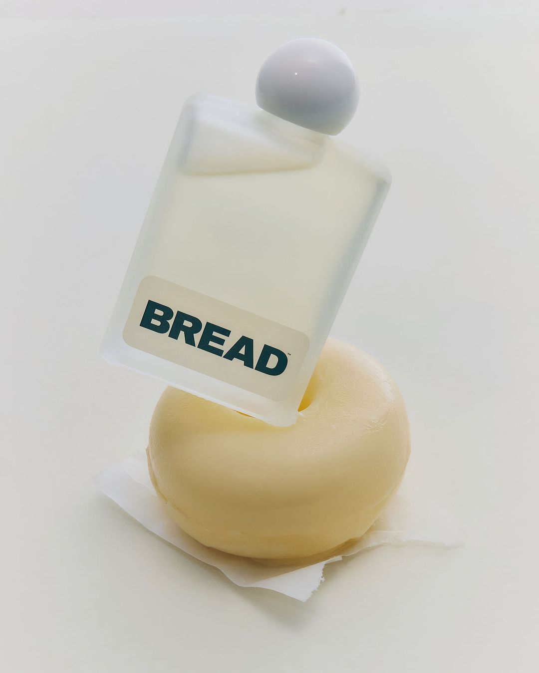 Still life of a frosted glass bottle with the word 'BREAD' displayed on it resting on a white donut and paper towel against a white background.