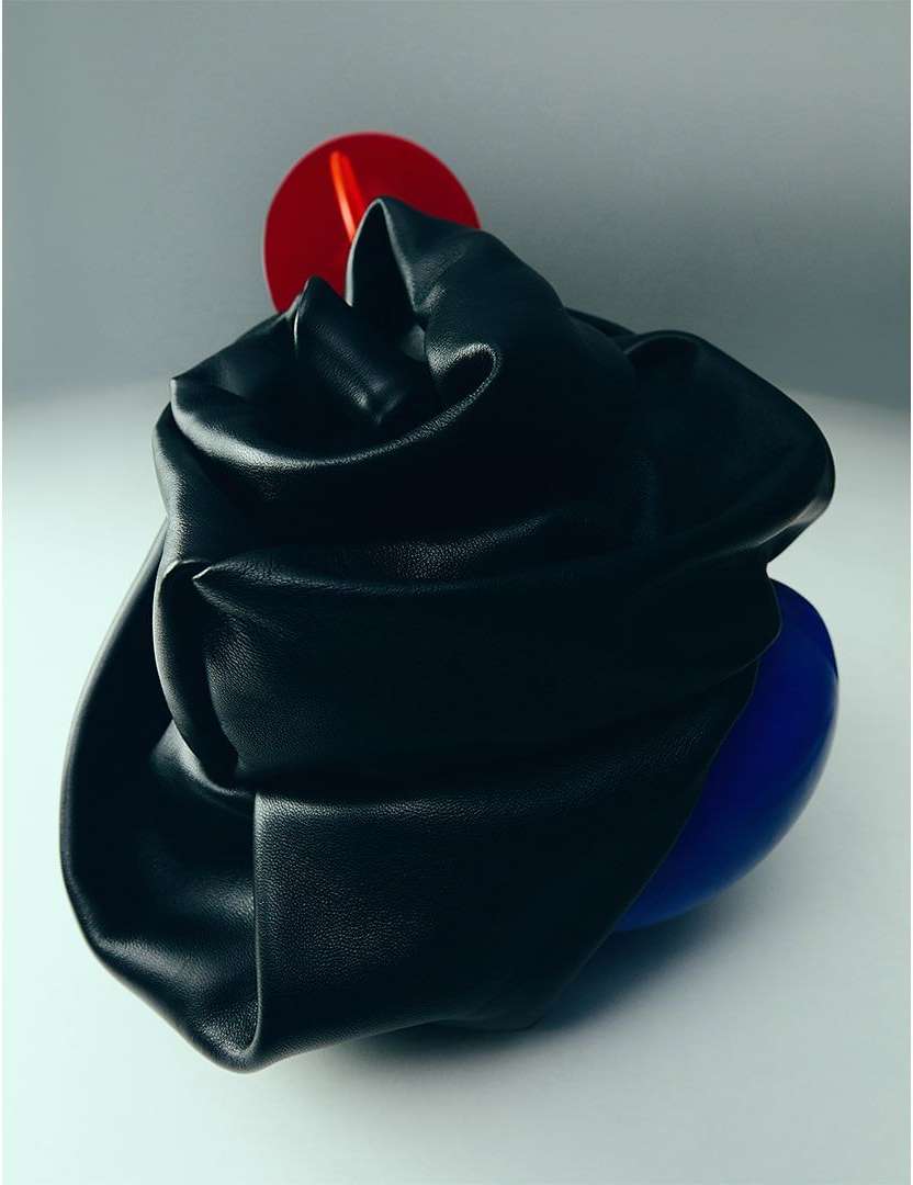 Abstract studio shot of a black leather object with draped form, accented by a red disc and a blue orb against a grey background.