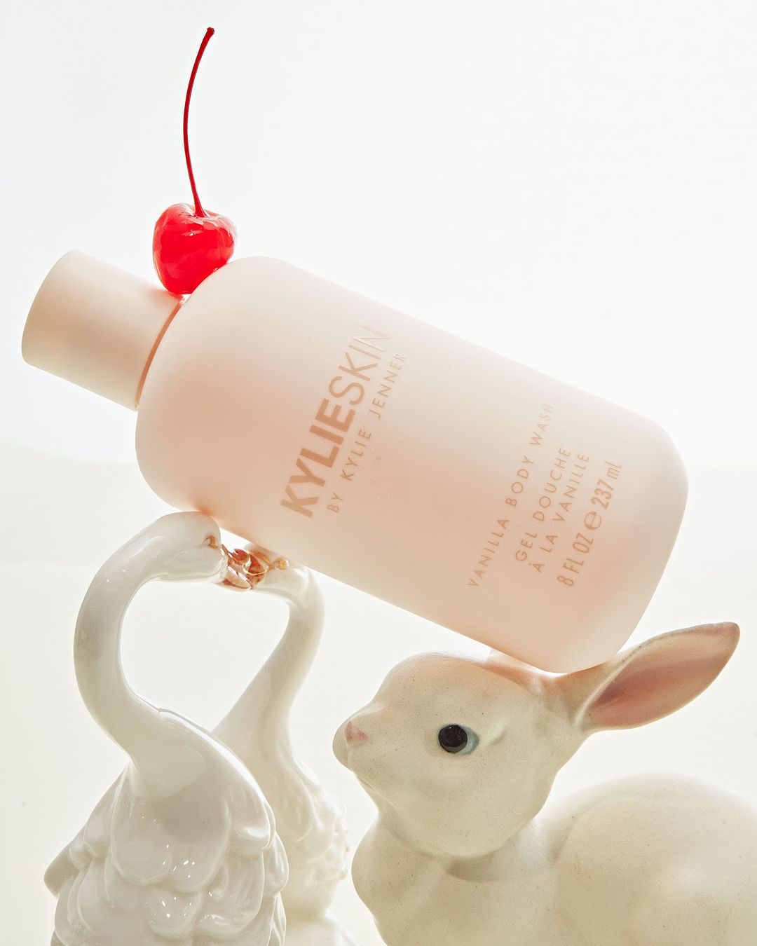 Kylie Skin Vanilla Body Wash bottle atop a white porcelain bunny and swan figurine set against a bright white background.