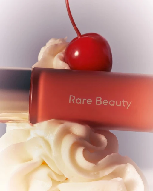 Rare Beauty product sits on whipped cream with a bright red cherry on top against a soft white background for a beauty product shot.