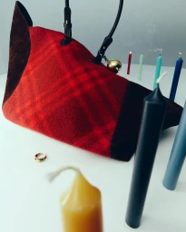 Red plaid handbag with leather straps and gold bell charm set among colorful lit candles on a white surface by Anna Pogossova.