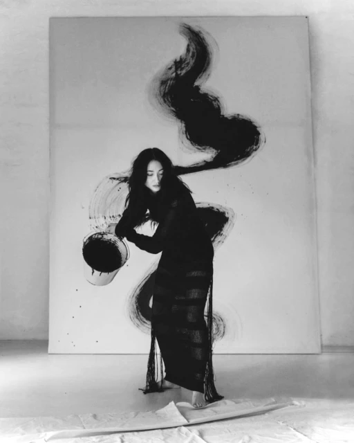 Monochrome image of a young woman in a long black dress, holding a bucket and painting abstract brushstrokes on a large white canvas.