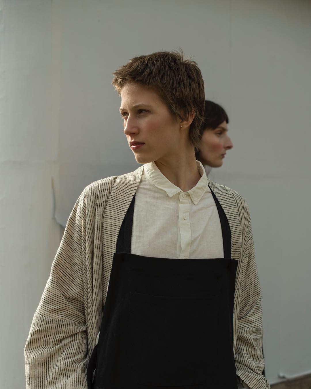 Two people with short hair pose in an indoor setting, wearing a black apron and striped jacket, in a medium portrait shot with soft lighting.