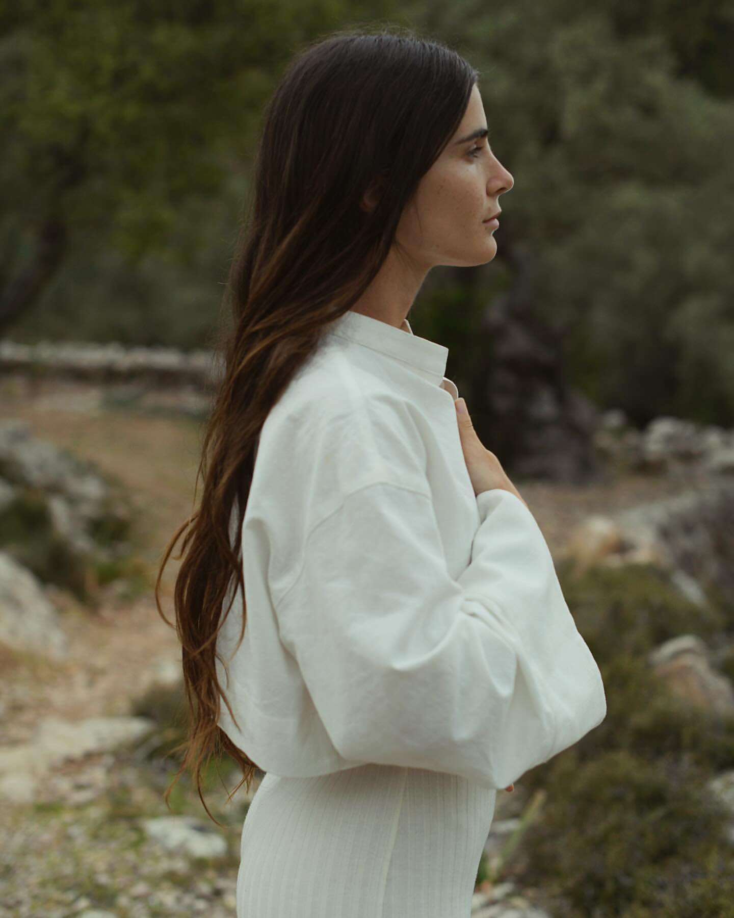 Woman in white dress poses outdoors, looking right in nature setting with neutral tones and soft light, captured in a medium shot.
