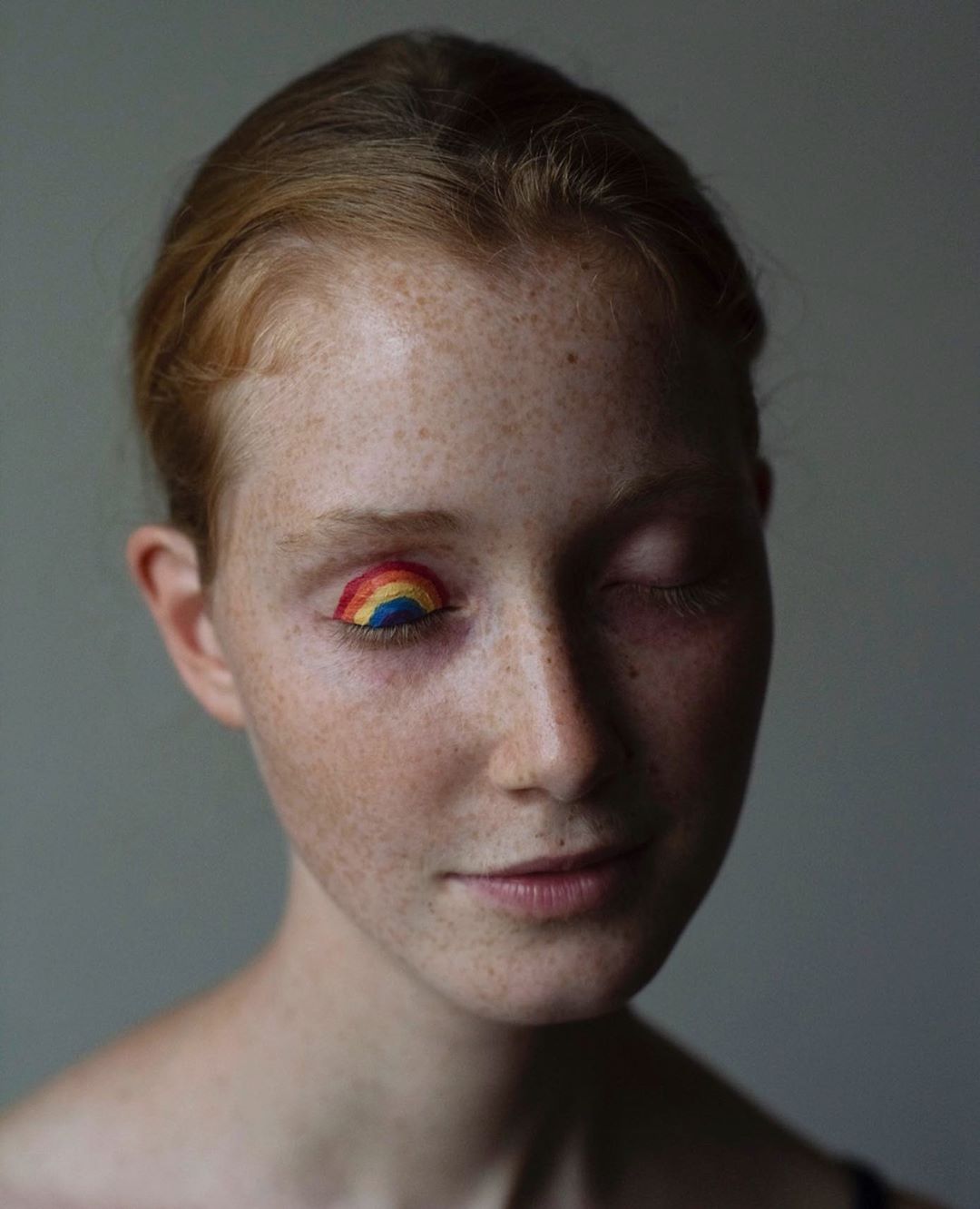Close-up portrait of a red-haired adult female with freckles, wearing rainbow colored eyeshadow on one closed eye, against a grey background.